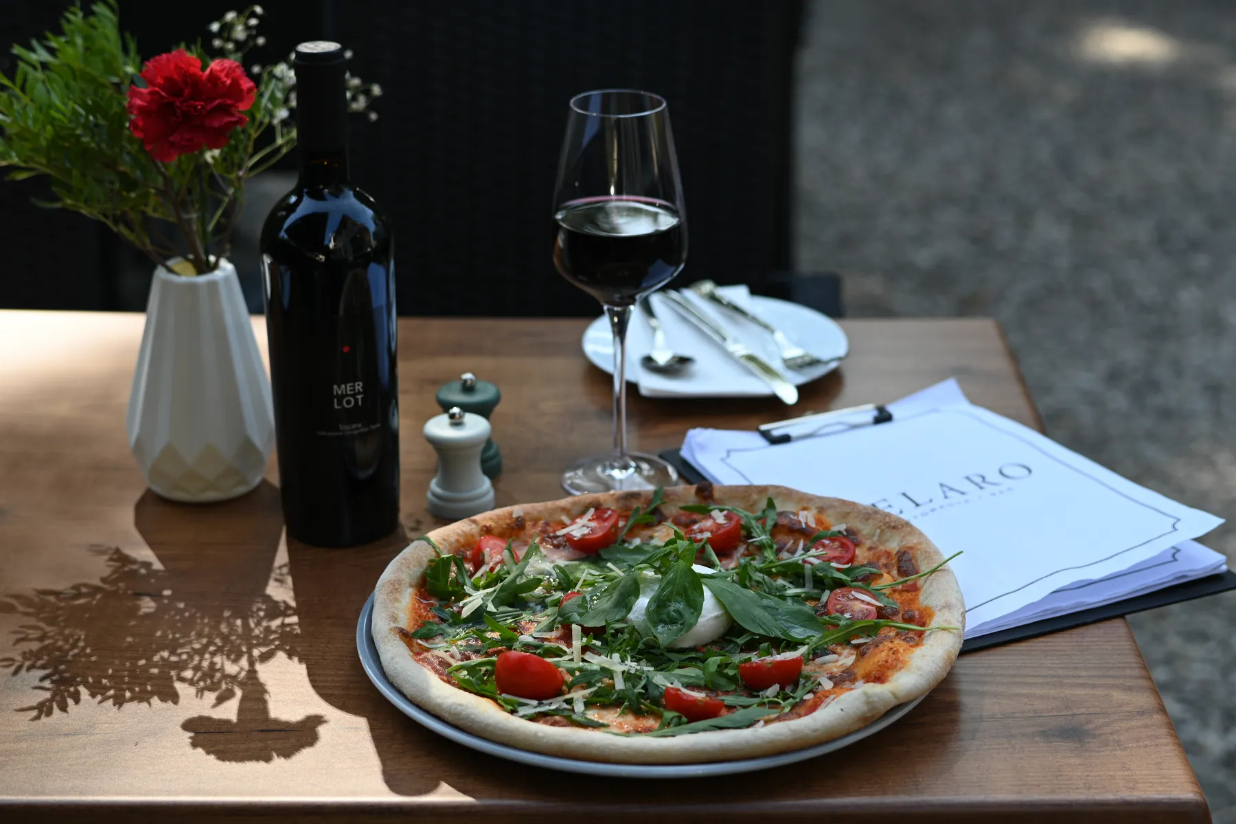 Ein Tisch auf der Terrasse: Pizza, Rotwein, rote Nelken und die VELARO-Speisekarte auf der Holzplatte